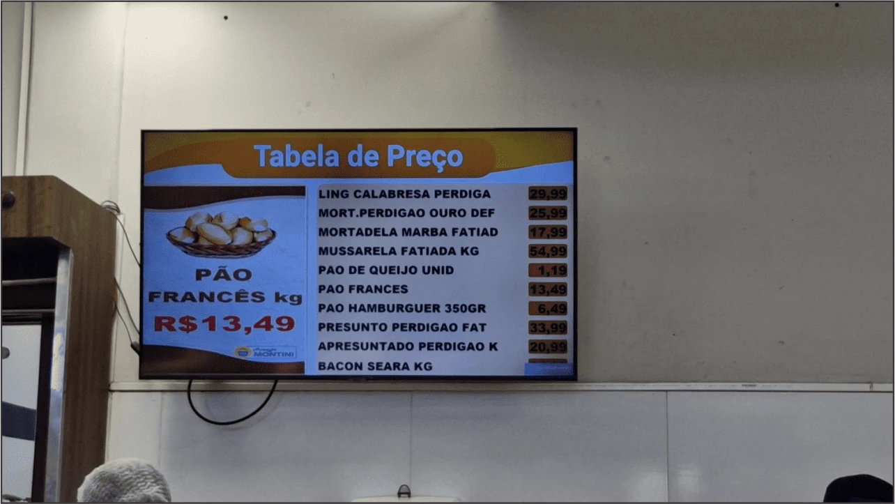Televisão exibindo tabela de preços de padaria e, ao lado, mídia promocional de pão francês